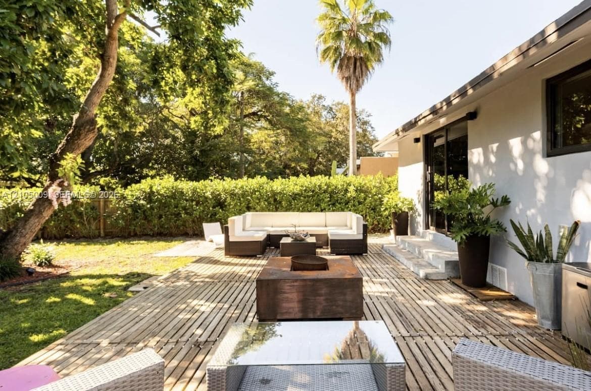 Expansive backyard featuring a wood-slat patio, a manicured lawn, lush perimeter hedging, and a white stucco exterior with dark-framed sliding glass doors