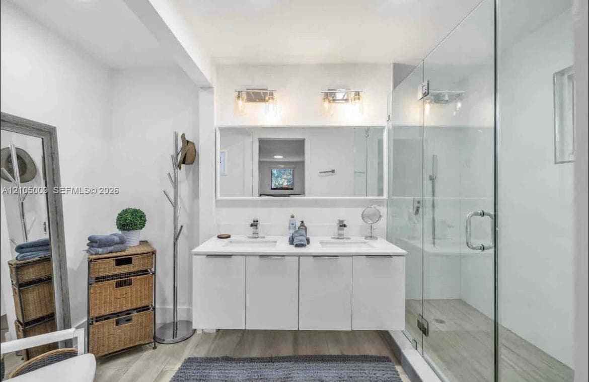 Dual sink vanity with white cabinetry, glass-enclosed shower, light wood-finish flooring, and contemporary wall-mounted light fixtures