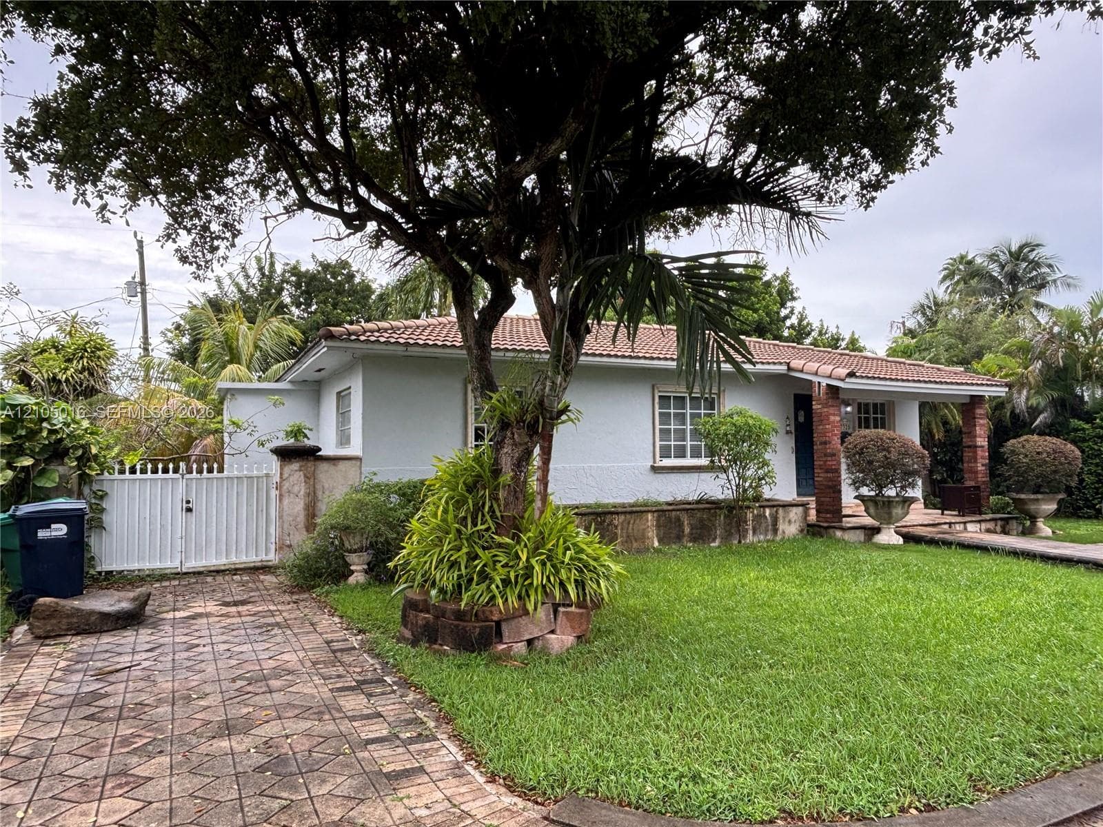 Stucco exterior with a terracotta tile roof, paver driveway, white gate, and mature trees