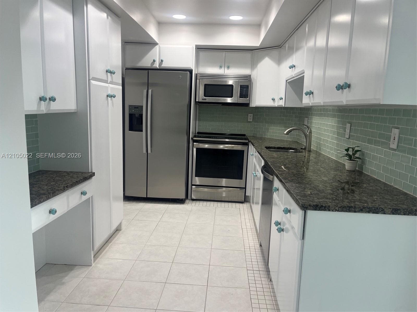 White cabinetry, dark stone countertops, stainless steel appliances, light tile flooring, and a green subway tile backsplash