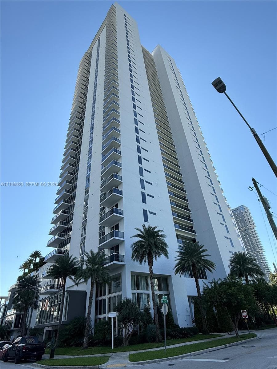 High-rise residential tower featuring a white concrete facade, vertical glass window panels, and integrated private balconies on each level, complemented by manicured palm tree landscaping