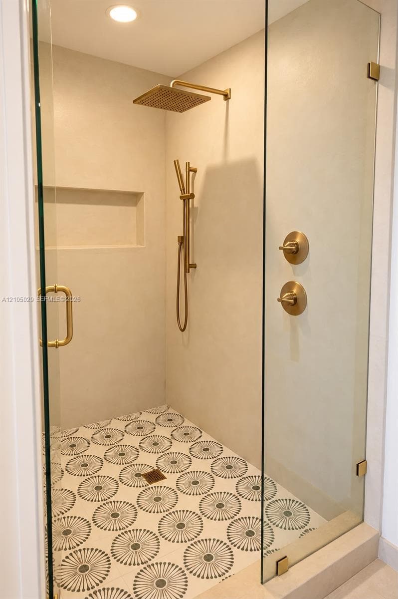 Walk-in shower featuring a frameless glass enclosure, patterned tile flooring, built-in wall niche, and brushed gold finish rainfall showerhead with matching handheld sprayer and controls