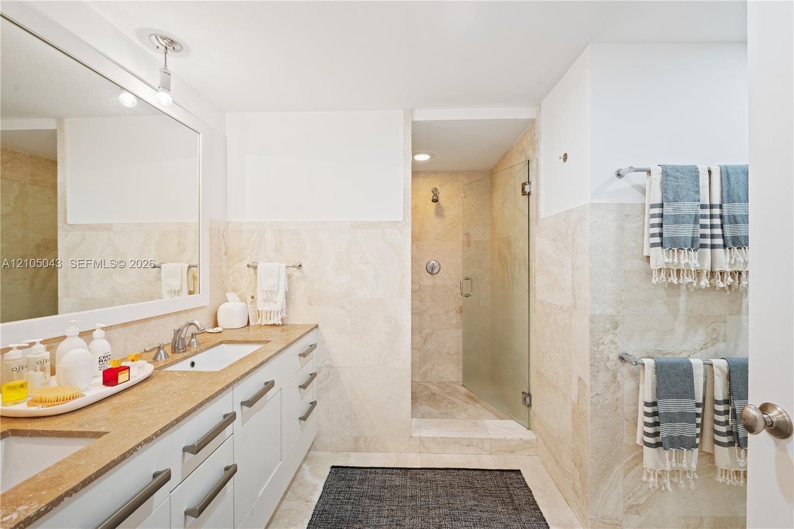 Dual-sink vanity with stone countertop, large framed wall mirror, stone-tiled shower enclosure with glass door, and wall-mounted metal towel bars