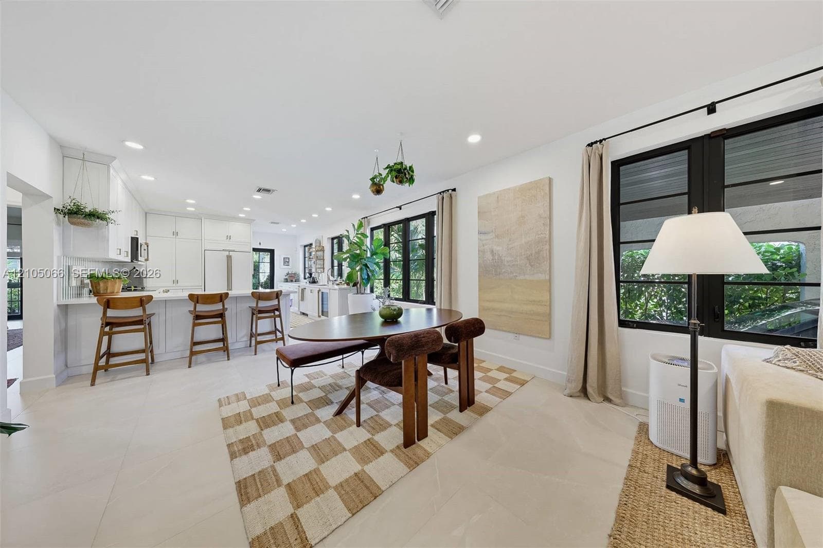 Open-concept kitchen and dining area featuring white shaker-style cabinetry, a central island with bar seating, large-format tile flooring, and oversized windows framed in black trim