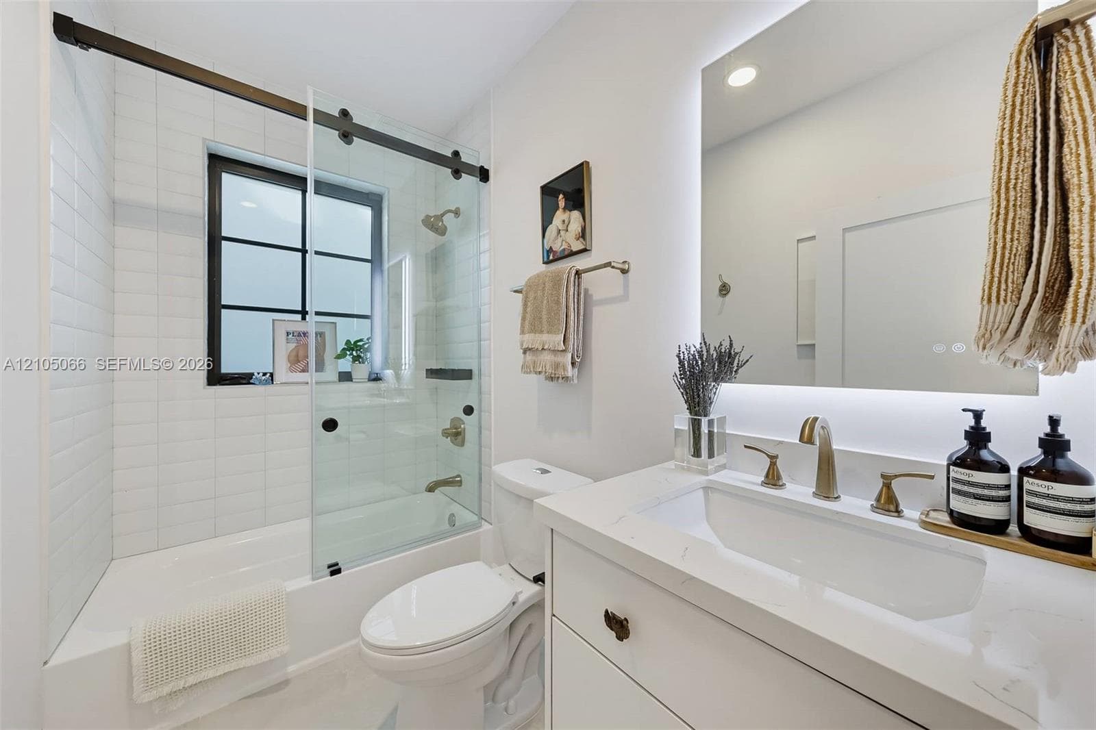 Bathroom featuring a vanity with a white countertop and brass-finish widespread faucet, a backlit mirror, a tub-shower combination with a sliding glass door and black hardware, and subway-style wall tile