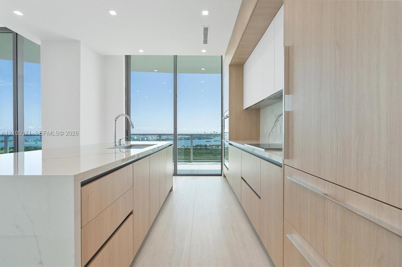Modern kitchen featuring wood-finish cabinetry, white matte upper cabinets, a waterfall-edge stone island, an integrated cooktop, and floor-to-ceiling glass sliding doors