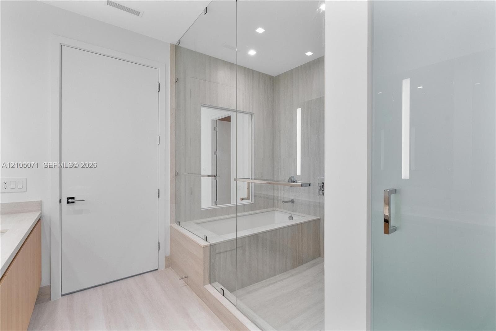 Glass-enclosed soaking tub, wall-to-wall neutral tile surround, recessed lighting, wood-finish vanity cabinetry, and frosted glass door