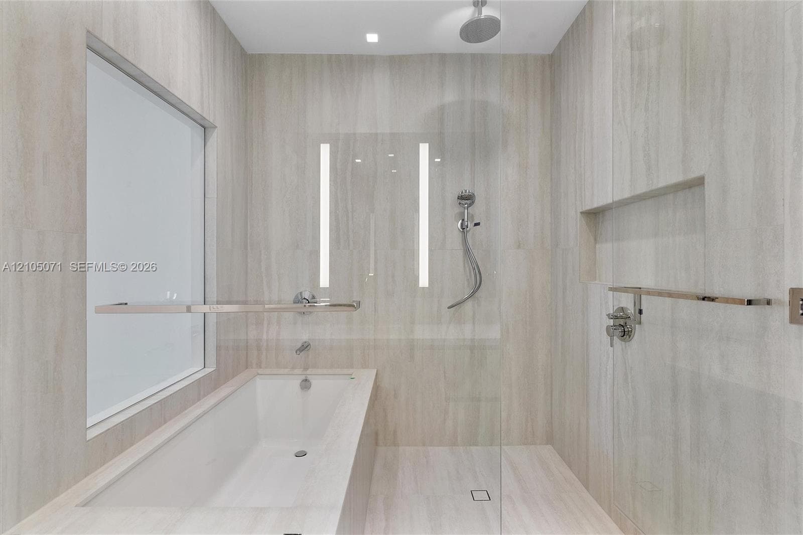 Full-height neutral stone-tile surround, soaking tub with polished chrome fixtures, glass-enclosed shower featuring a rainfall showerhead, and integrated wall niche