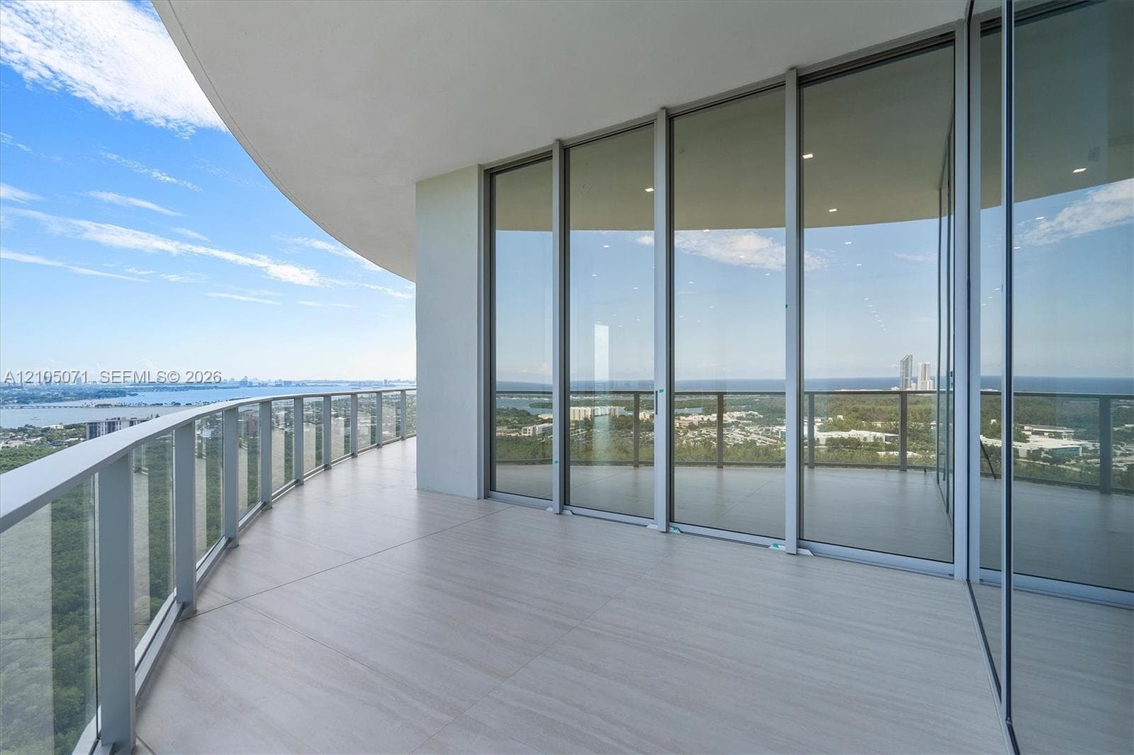 Curved covered balcony featuring large-format porcelain tile flooring, expansive floor-to-ceiling glass sliding doors, and a transparent tempered glass railing