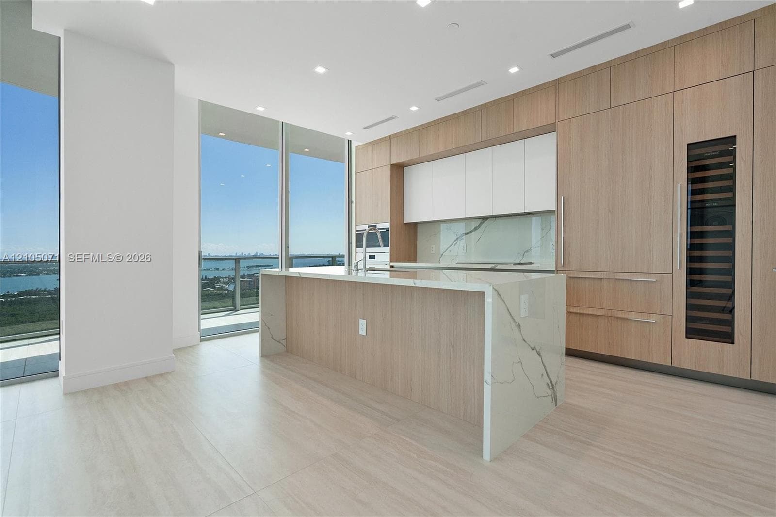 Contemporary kitchen featuring light wood-finish cabinetry, a waterfall-edge stone island, integrated refrigeration, and floor-to-ceiling glass paneling