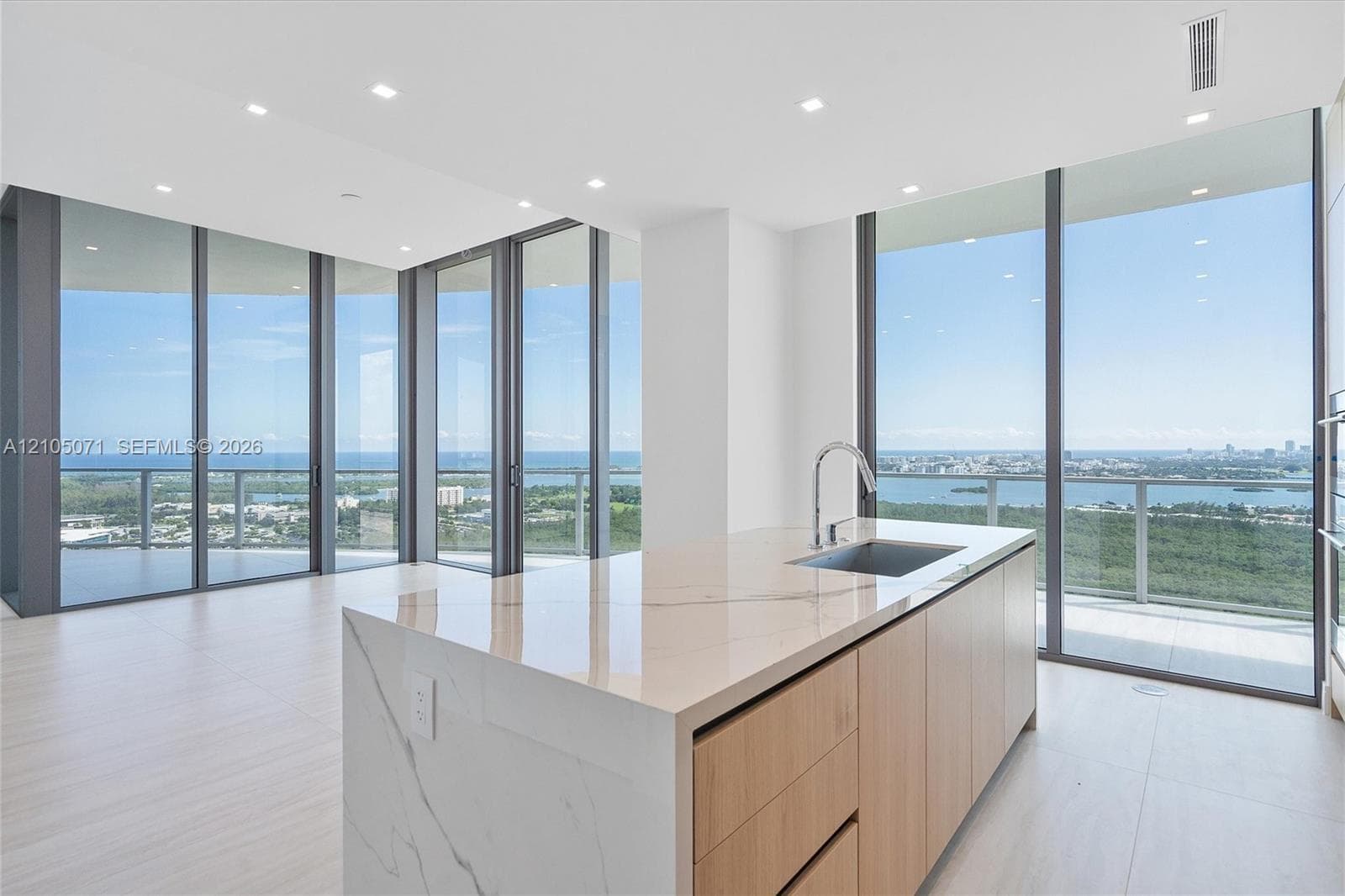 Kitchen island featuring a stone waterfall countertop, integrated undermount sink with a gooseneck faucet, light wood-finish cabinetry, floor-to-ceiling glass panel walls, and recessed lighting