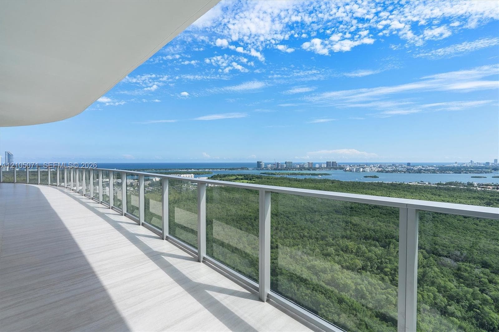 Expansive wraparound balcony featuring neutral wood-finish flooring, a glass panel railing, and an overhead architectural canopy