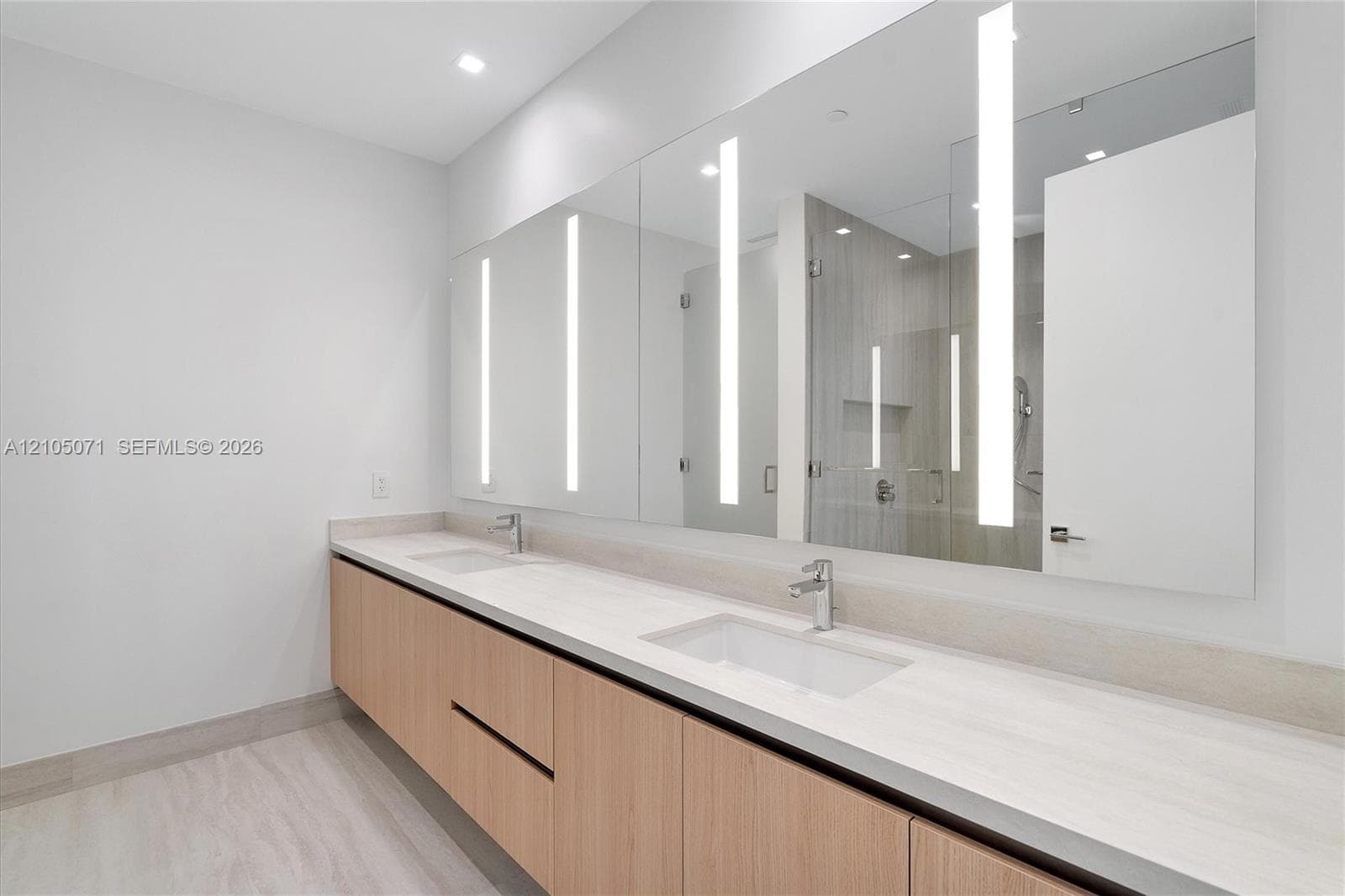 Floating vanity with light wood-finish cabinetry, dual rectangular undermount sinks, stone-surface countertop, and a full-width mirror featuring integrated vertical lighting