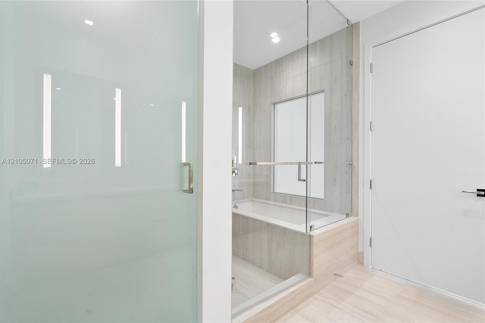 Bathroom featuring a glass-enclosed shower, a built-in soaking tub with light-toned tile surround, neutral tile flooring, and integrated wall lighting