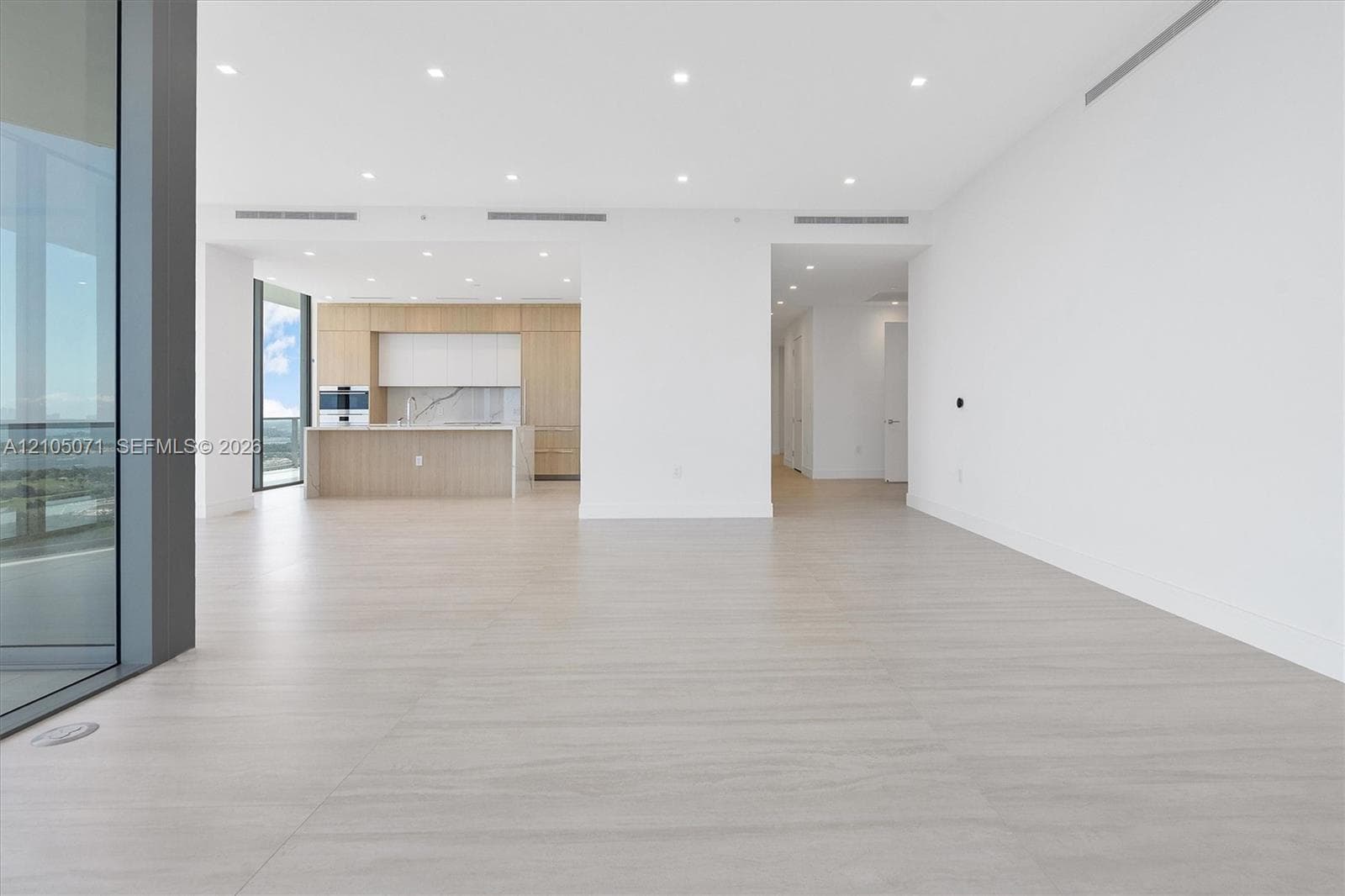 Open-concept interior featuring light-toned wood-finish flooring, recessed ceiling lighting, floor-to-ceiling glass paneling, and a kitchen equipped with built-in cabinetry and a stone-surface island