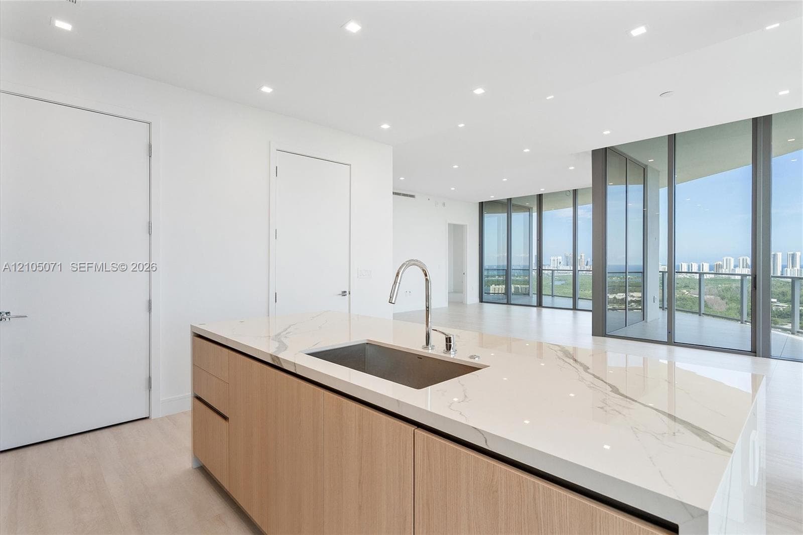 Kitchen island featuring a veined stone countertop, stainless steel undermount sink, high-arc faucet, and light wood-finish cabinetry with recessed lighting and floor-to-ceiling glass windows