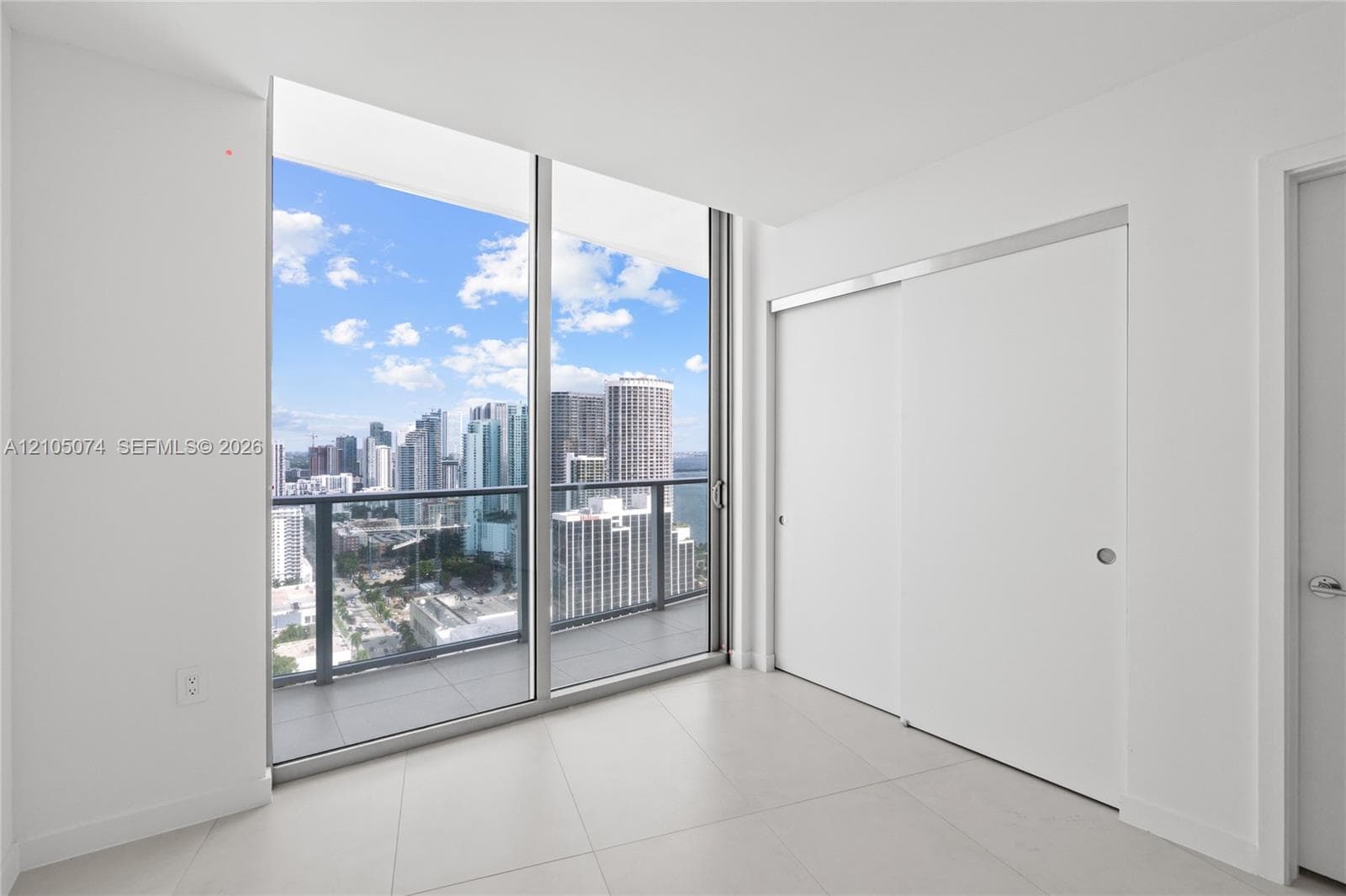 Floor-to-ceiling glass sliding doors, private balcony with metal railing, light-colored tile flooring, and urban horizon scenery