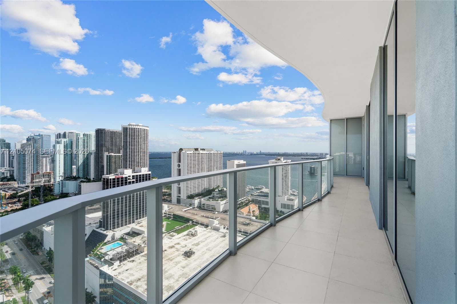 Curved exterior balcony featuring large-format tile flooring, a glass panel railing with a dark metal frame, and sliding glass door access