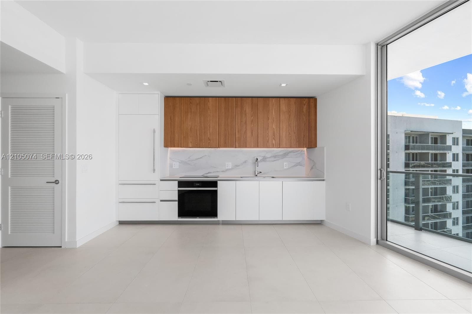 Modern kitchen featuring slab-front wood cabinetry, white stone-finish countertops, full-height stone-finish backsplash, built-in cooktop, and floor-to-ceiling windows with balcony access