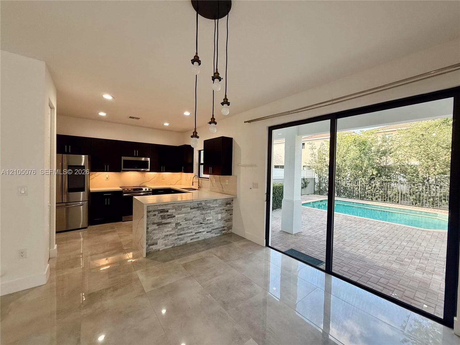 Kitchen featuring dark cabinetry, stone-veneer peninsula, recessed lighting, and large-format tile flooring, accompanied by expansive sliding glass doors opening to a paved patio and swimming pool