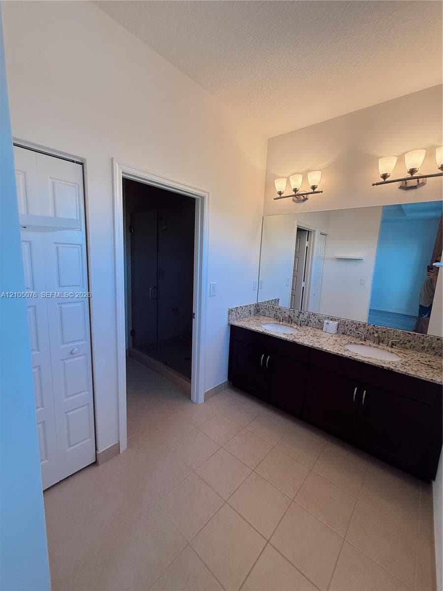 Bathroom featuring a dual-sink vanity with stone-finish countertop, dark cabinetry, large wall-mounted mirror, and tile flooring