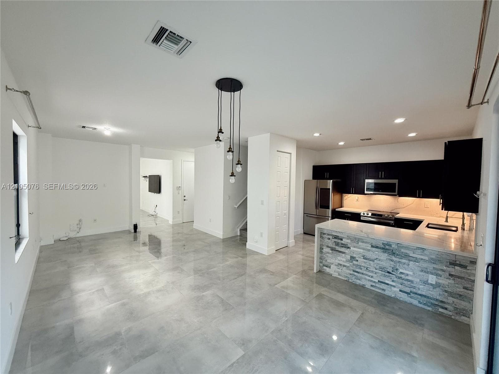 Open-concept area featuring oversized neutral tile flooring, kitchen with dark cabinetry, stone-veneer breakfast bar, and industrial-style pendant lighting