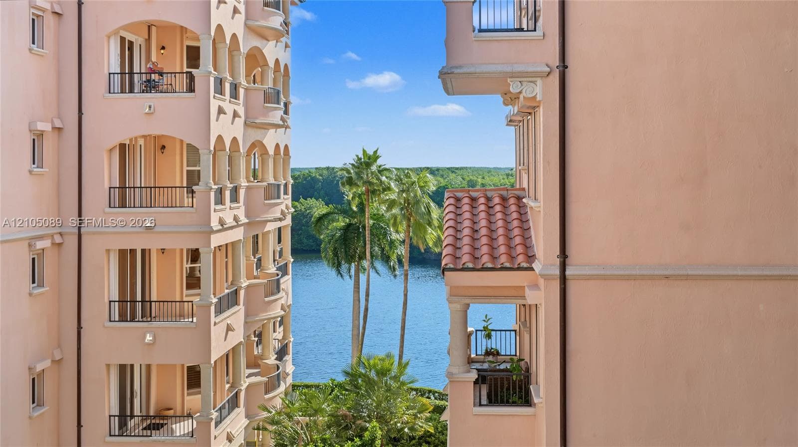 Stucco exterior with multi-level arched balconies, metal railings, column supports, and clay tile roof accents, featuring a water backdrop with palm trees
