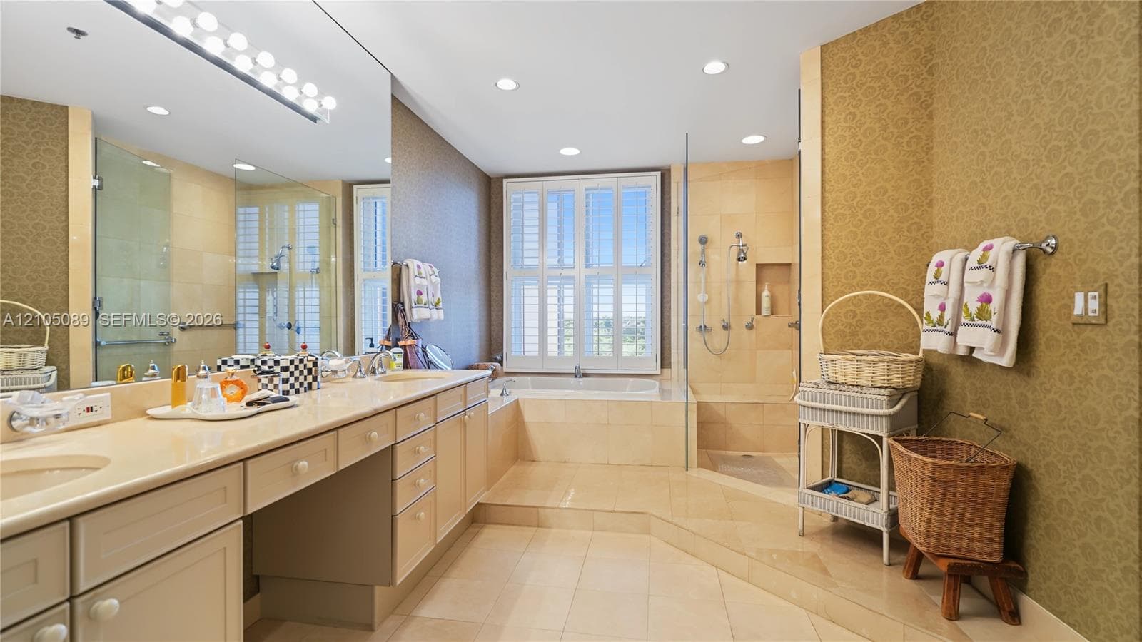 Extended vanity with dual integrated sinks and stone countertop, soaking tub encased in stone tile, walk-in glass-enclosed shower with multiple showerheads, and interior window shutters