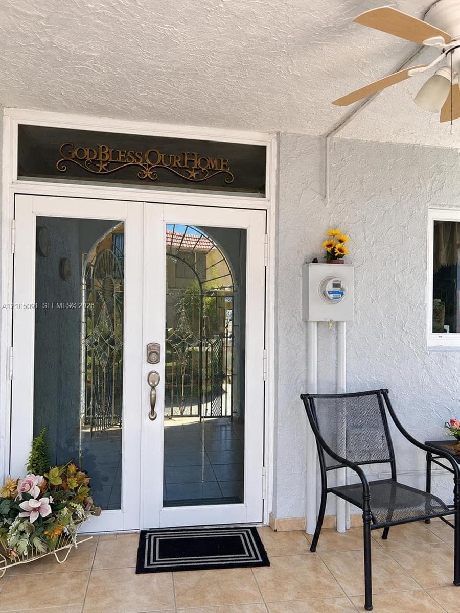 Double entry doors with glass inserts and transom window, paired with a covered porch featuring tiled flooring and a ceiling-mounted fan