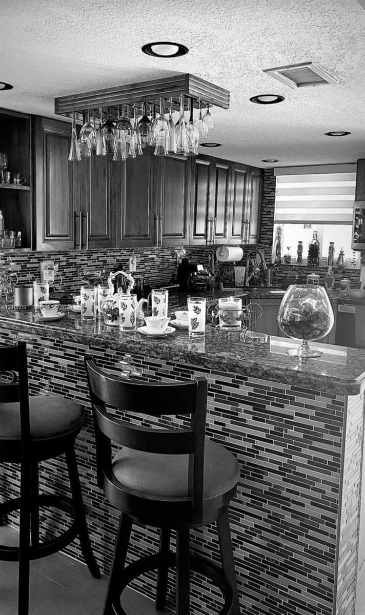 Kitchen featuring wood cabinetry, stone countertops, multi-toned linear tile backsplash, and a built-in suspended stemware rack