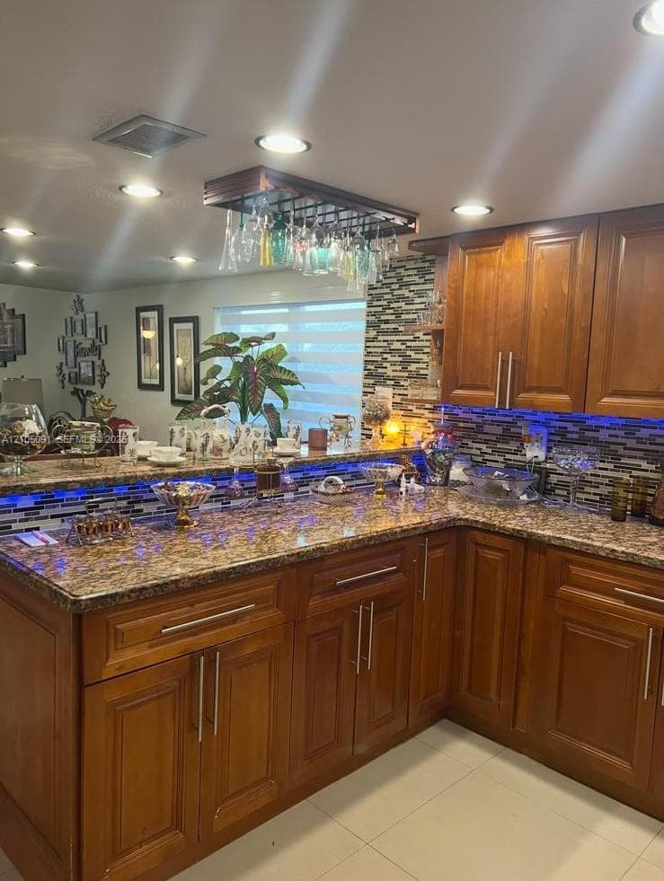 Kitchen featuring stained wood cabinetry, stone-patterned countertops, a decorative mosaic tile backsplash, and a built-in overhead glass rack