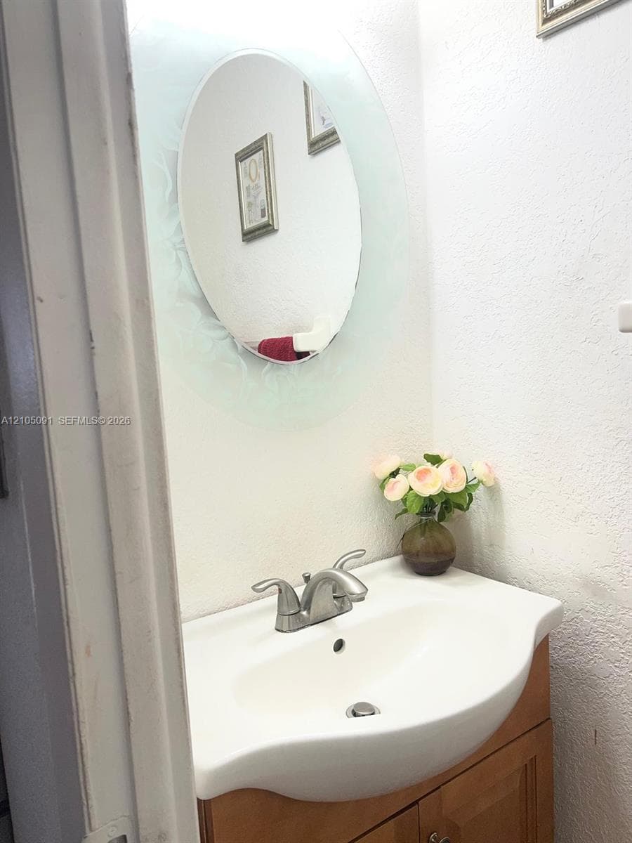 Vanity featuring a light-toned wood-finish cabinet, integrated ceramic sink basin, metal double-handle faucet, and an oval mirror with a decorative frosted glass border