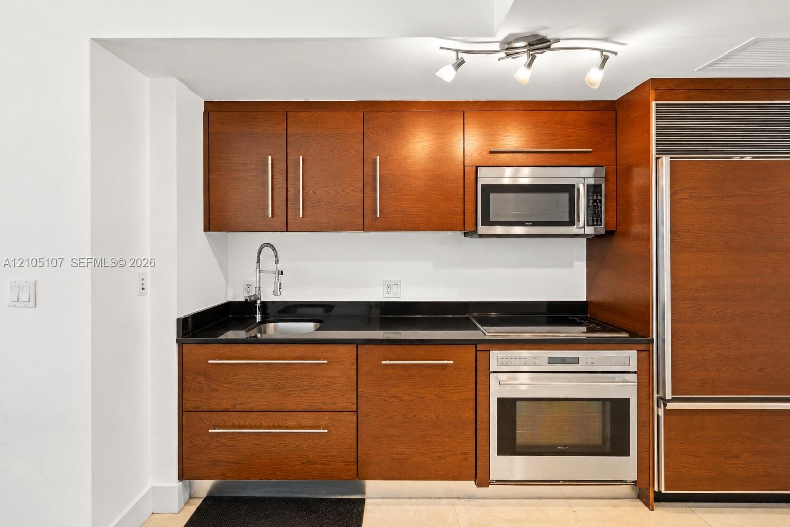 Kitchen featuring wood-finish cabinetry, integrated stainless steel appliances, black countertops, and a track lighting fixture