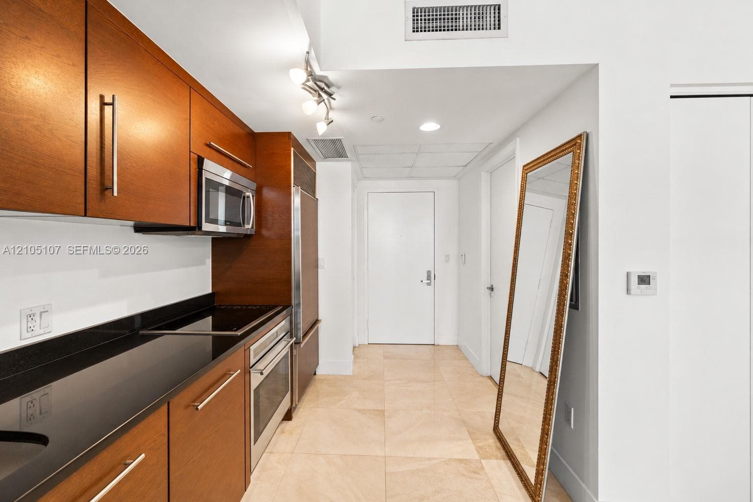 Galley kitchen with wood cabinetry, dark countertops, integrated stainless steel appliances, and tile flooring