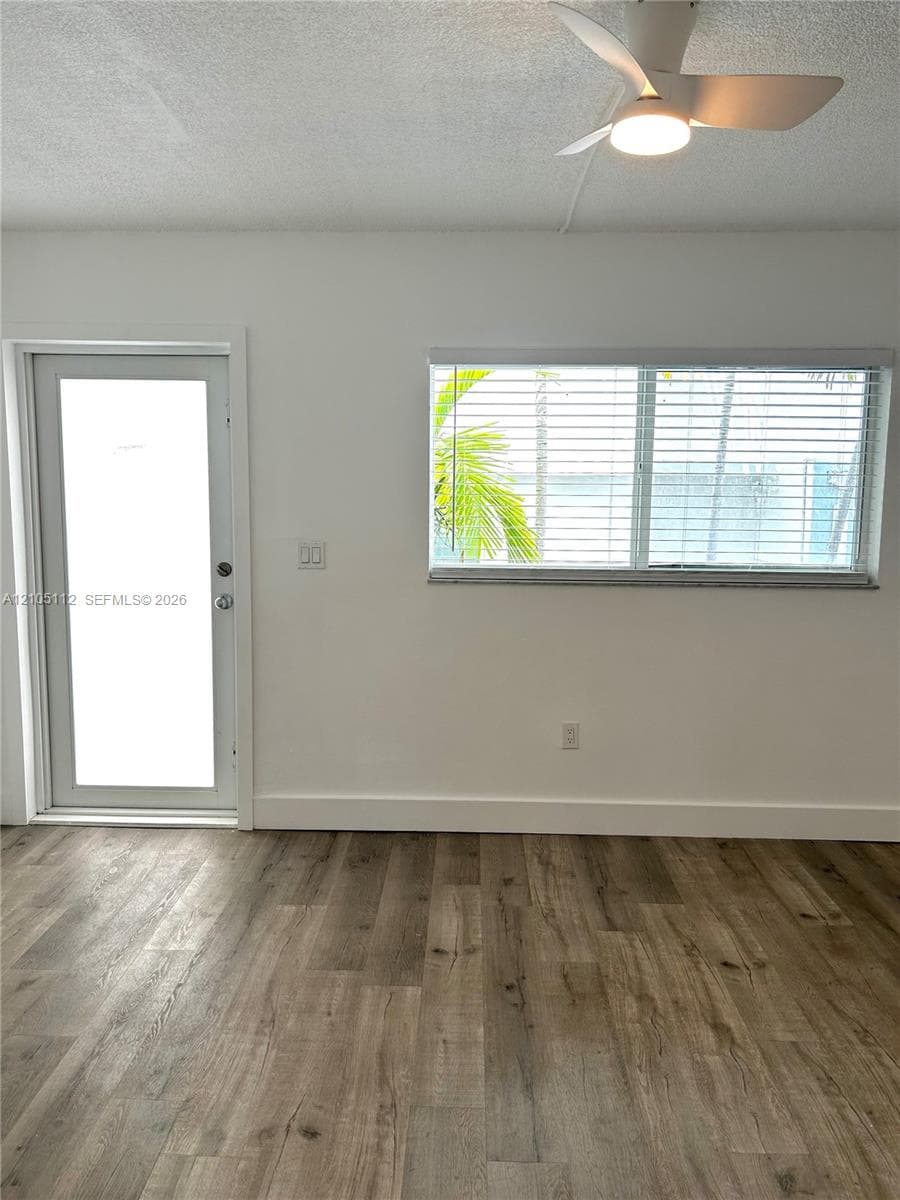 Wood-finish flooring, white-framed exterior door with glass pane, large window with horizontal blinds, and a mounted modern ceiling fan