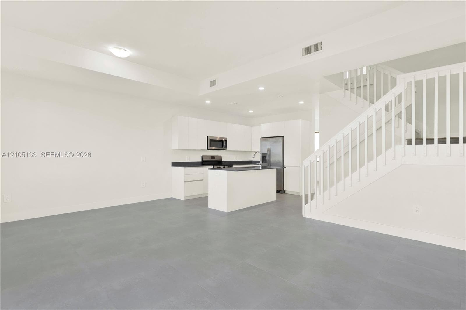 Open-concept main level featuring light gray flooring, a contemporary kitchen with white cabinetry and dark countertops, stainless steel appliances, and a staircase with white balusters