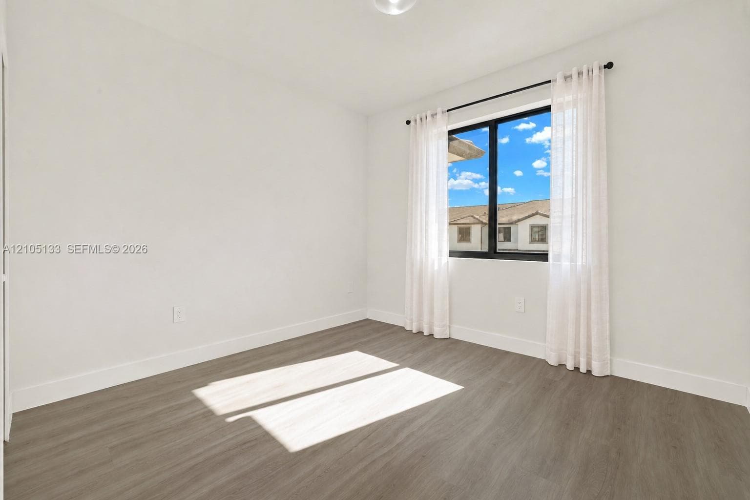 Spacious room featuring wood-finish flooring, white walls, and a large window with sheers