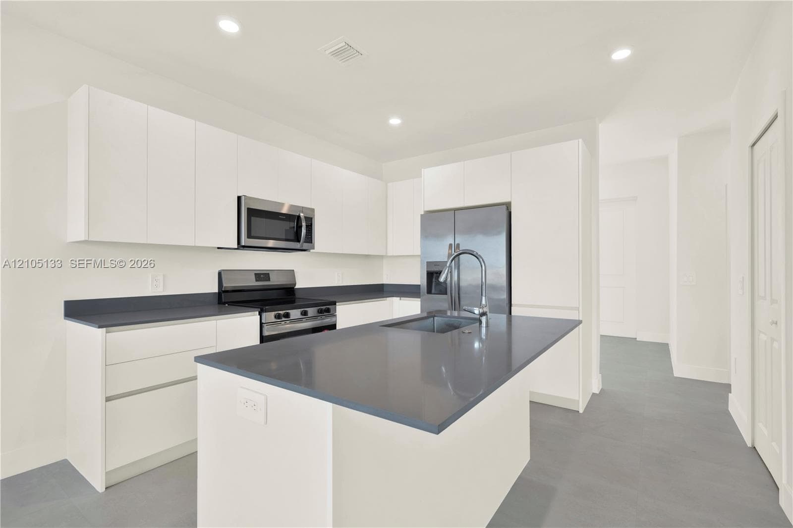 Modern kitchen featuring white flat-panel cabinetry, stainless steel appliances, dark countertops, a central island with an integrated sink, and recessed lighting