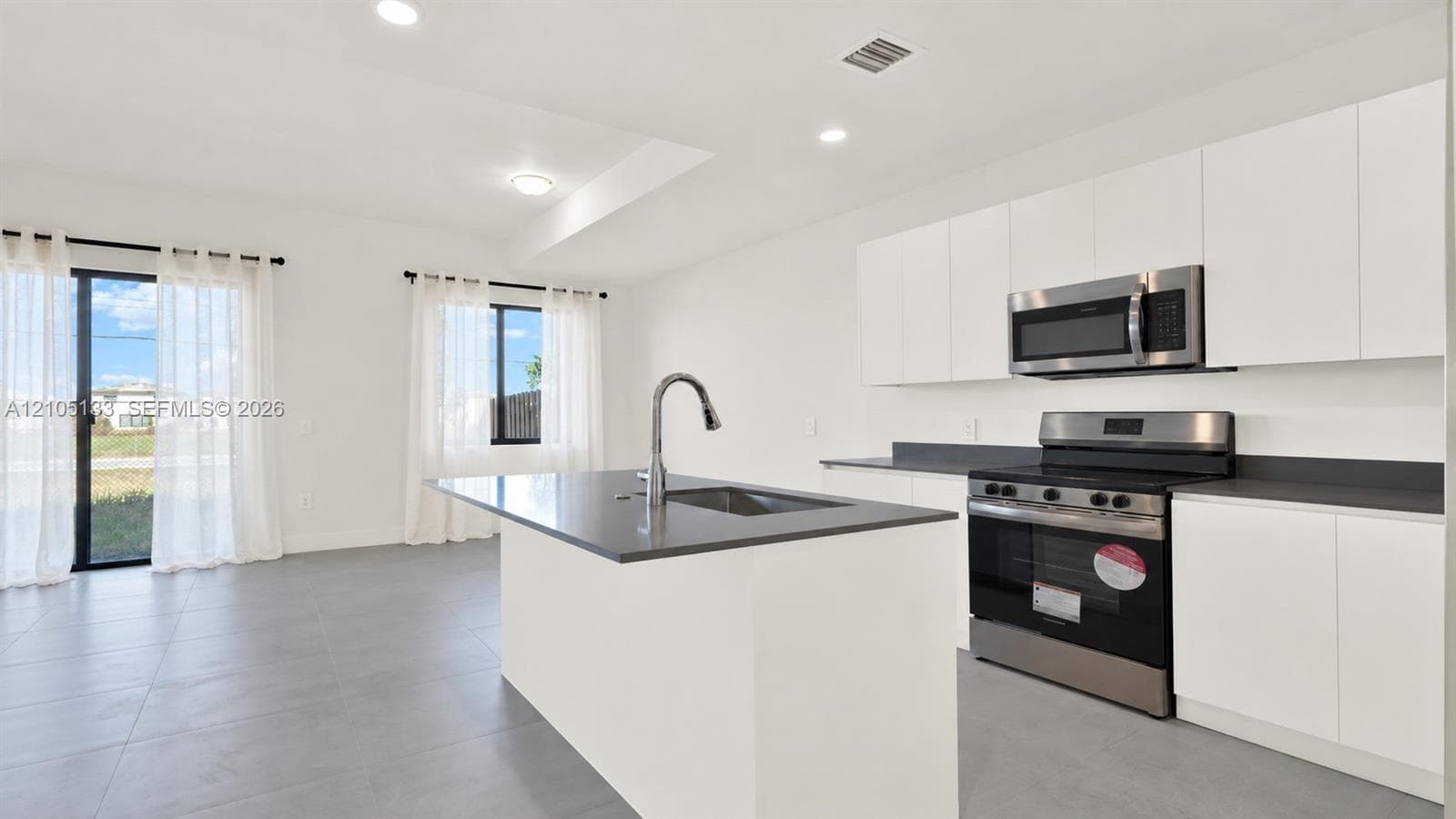 Modern kitchen featuring a stainless steel range and microwave, white flat-panel cabinetry, a kitchen island with an integrated sink, and dark countertops