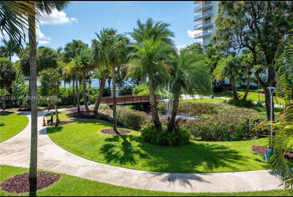 Landscaped grounds featuring mature palm trees, a concrete walking path, a wooden pedestrian bridge, and a central water feature with a fountain