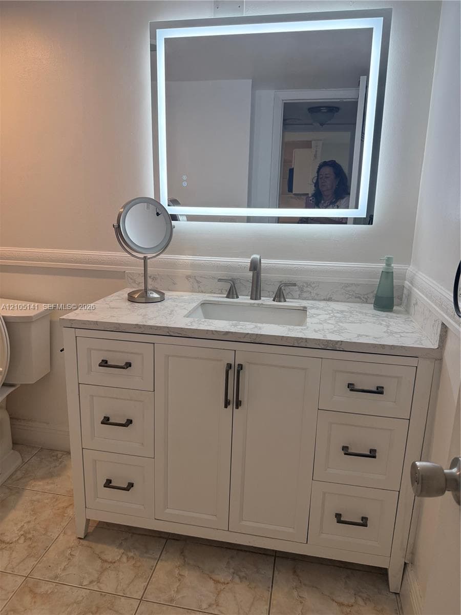 White vanity with stone-patterned countertop, integrated rectangular sink, matte-finish hardware, and wall-mounted illuminated rectangular mirror