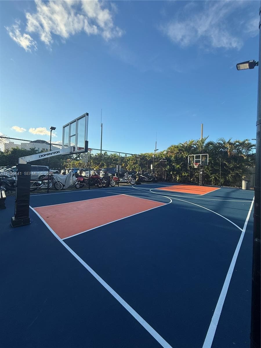 Outdoor basketball court featuring multi-colored hardcourt surfacing, perimeter fencing, dual hoop systems, and integrated overhead lighting