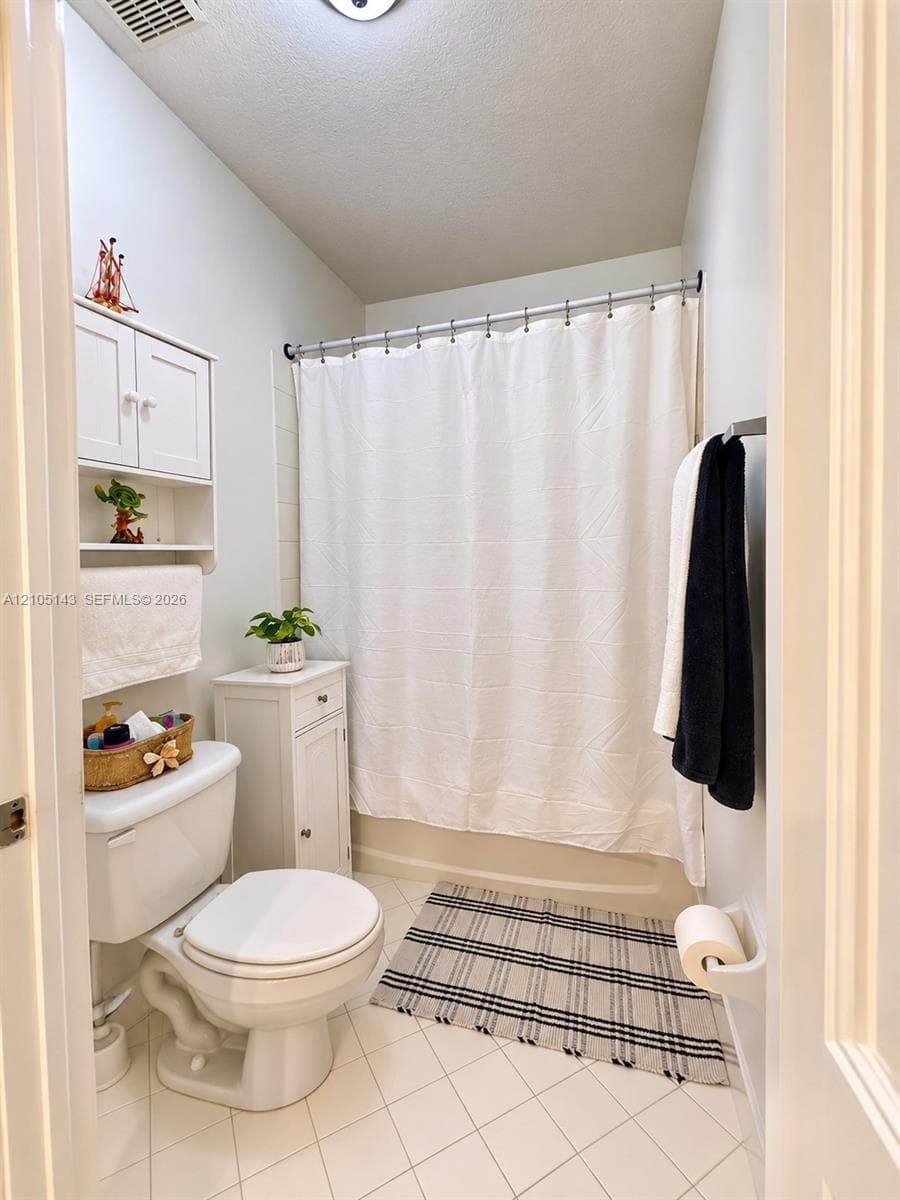 White tile flooring, a built-in toilet, a white wall-mounted storage cabinet, and a shower with a white curtain