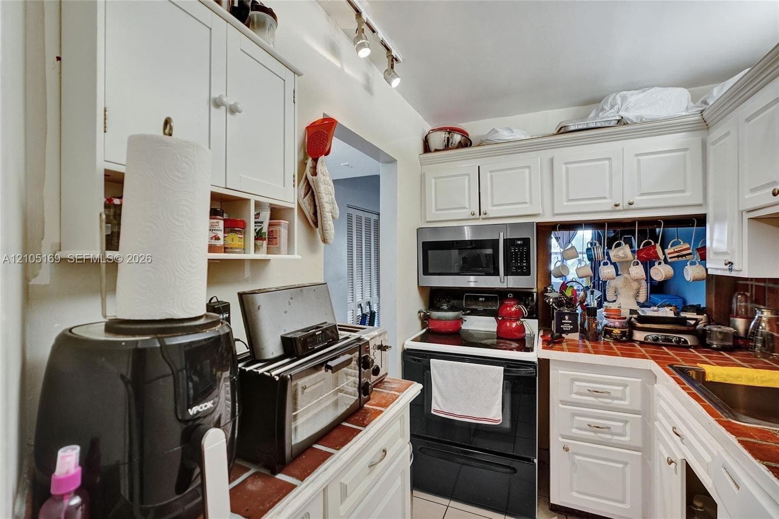 White cabinetry, stainless steel microwave, track lighting, tiled countertop, and stainless steel sink