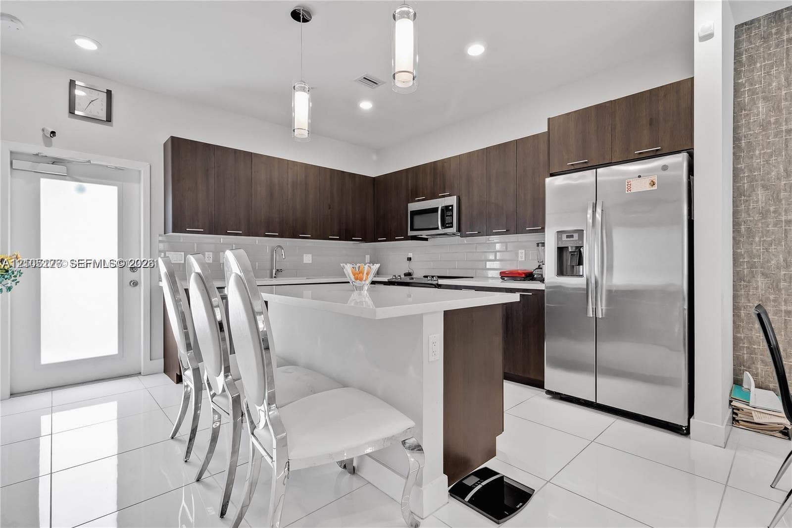 Modern kitchen featuring dark wood-finish cabinetry, a central island with white countertop seating, white tile flooring, stainless steel appliances, and minimalist pendant lighting
