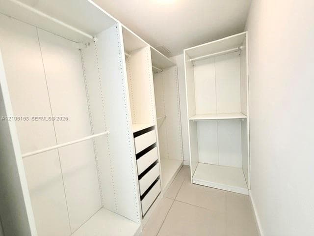 Walk-in closet featuring white custom built-in shelving, integrated hanging rods, and a four-drawer storage unit with large-format tile flooring
