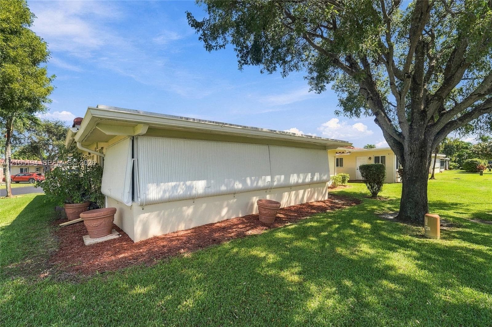 Stucco exterior featuring retractable storm shutters and a manicured lawn with mature shade trees