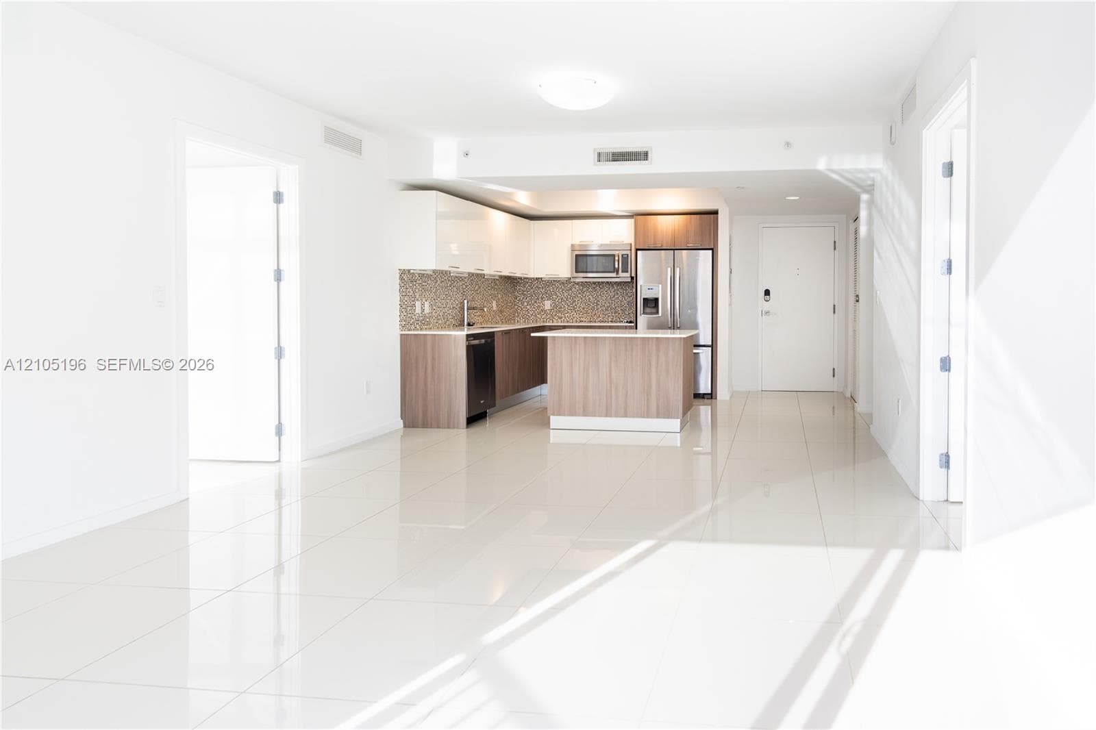Spacious open-concept living area with polished tile flooring, a kitchen island, integrated stainless steel appliances, wood-finish cabinetry, and a mosaic tile backsplash