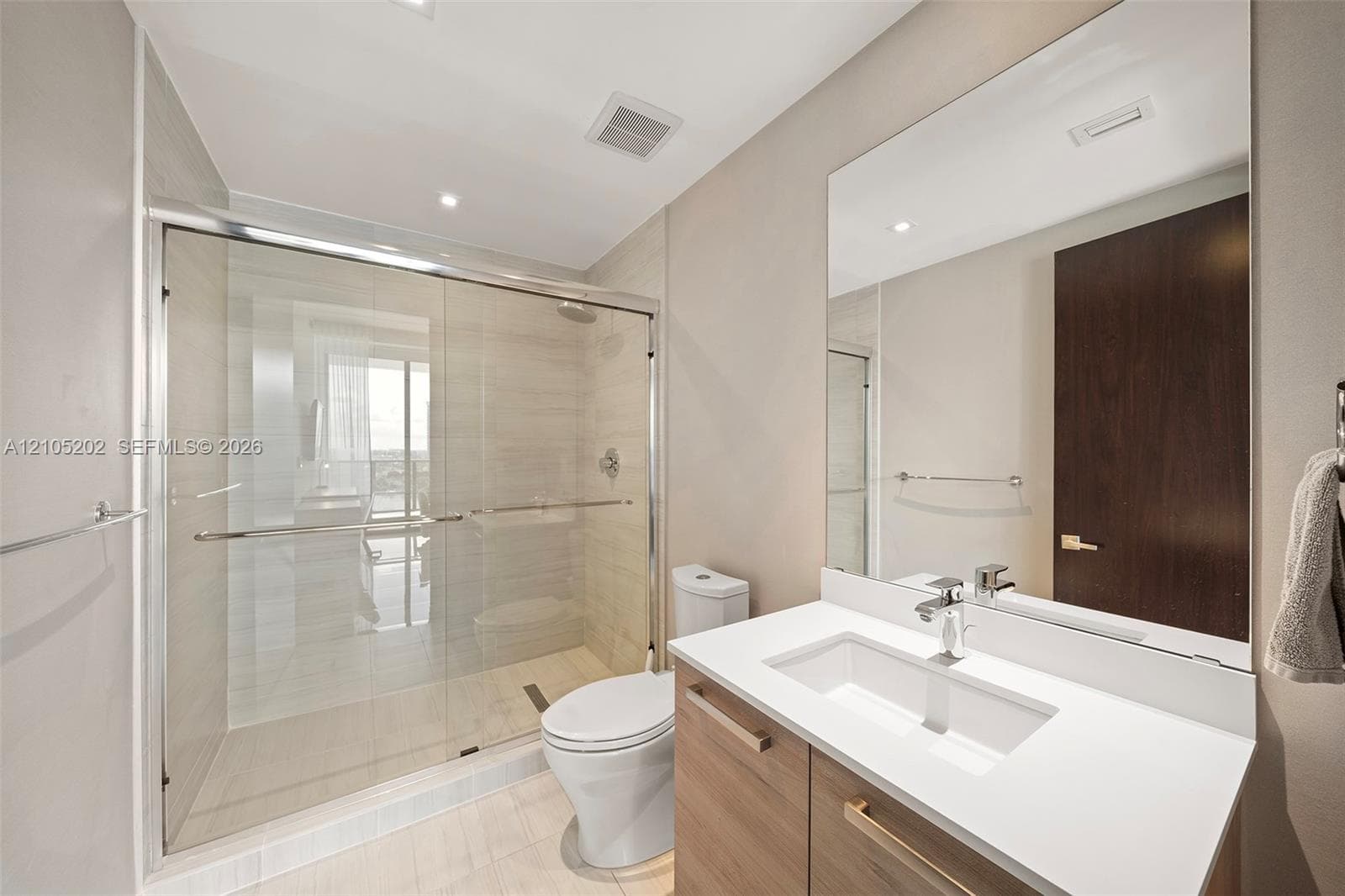 Bathroom featuring a walk-in shower with glass enclosure, light-tone tile surround, wood-finish vanity with stone-style countertop, rectangular undermount sink, and full-width vanity mirror
