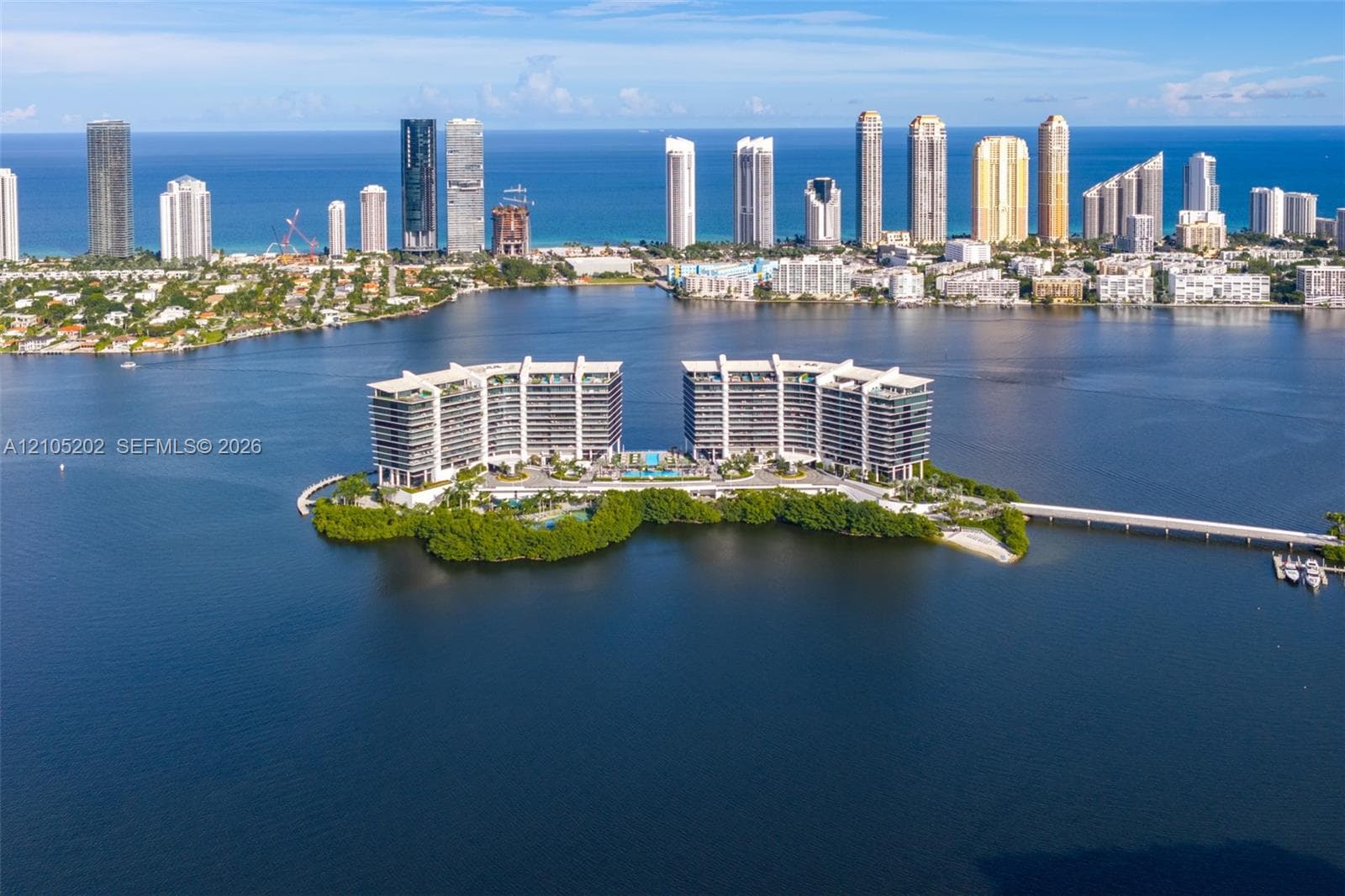 Aerial perspective of an island property featuring waterfront positioning, dual mid-rise structures, a private causeway entrance, and extensive perimeter foliage